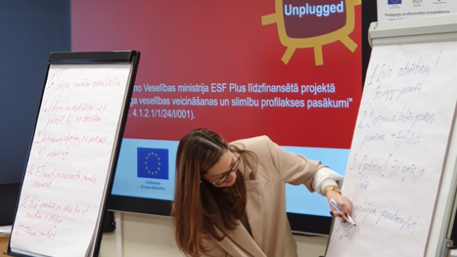 Cilvēks stāv pie papīra tāfeles un no tās lasa tekstu. Cilvēkam fonā LED ekrāns ar prezentācijas slaidu.