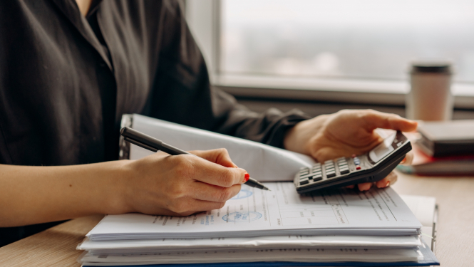 Attēlā pie galda sēž cilvēks ar dokumentu mapi un kalkulatoru 