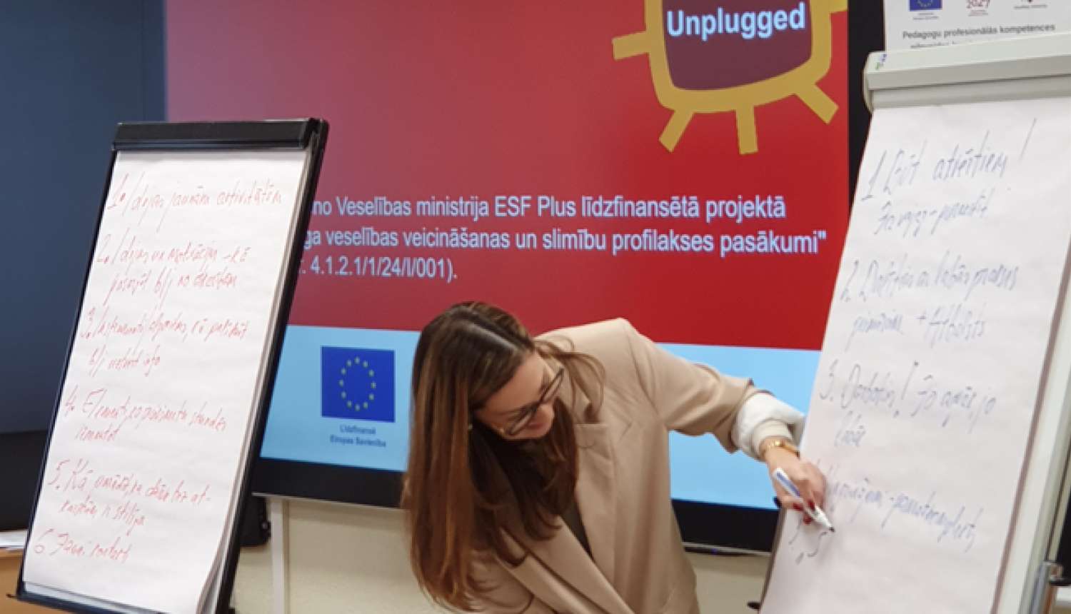 Cilvēks stāv pie papīra tāfeles un no tās lasa tekstu. Cilvēkam fonā LED ekrāns ar prezentācijas slaidu.