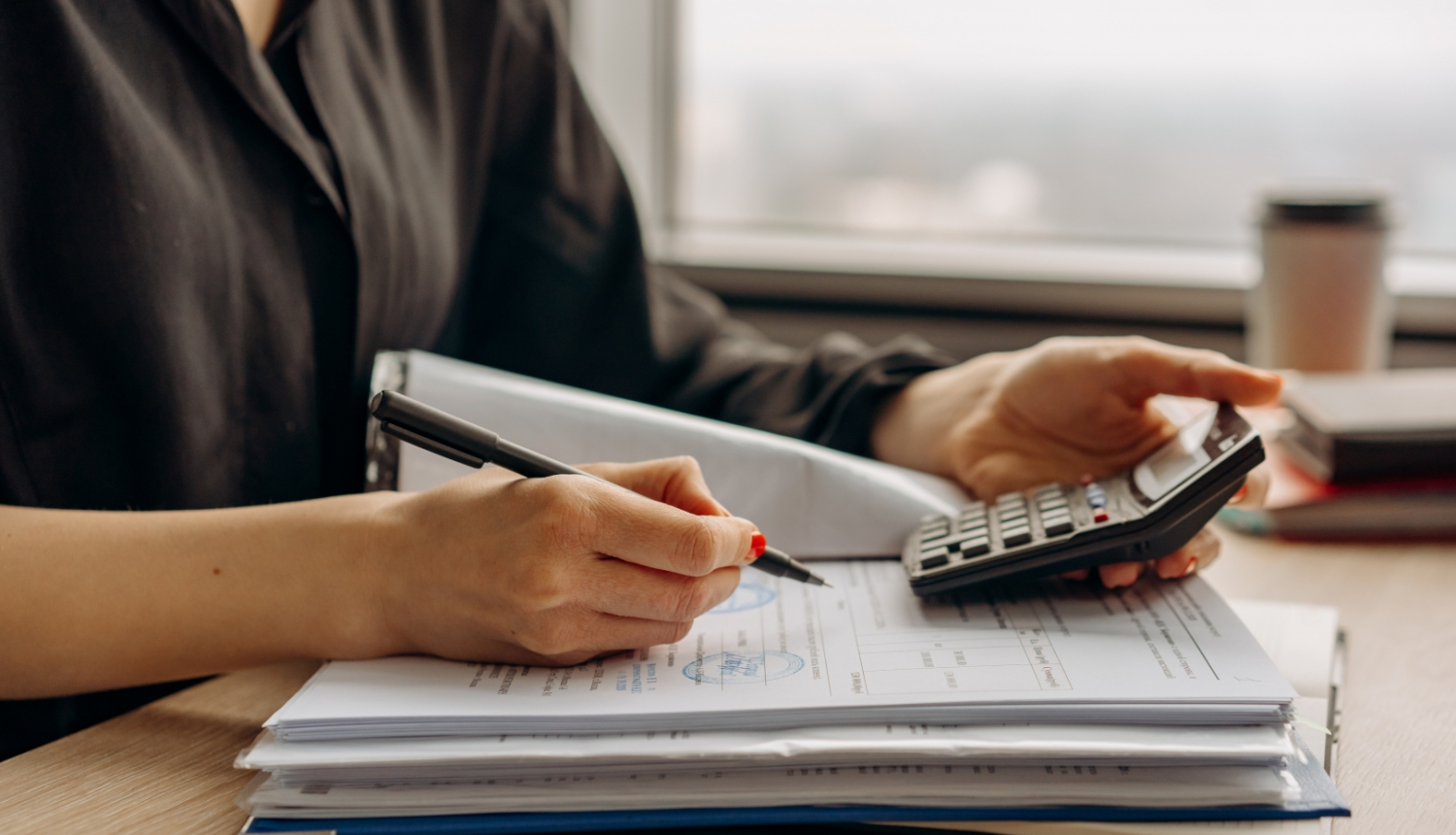 Attēlā pie galda sēž cilvēks ar dokumentu mapi un kalkulatoru 